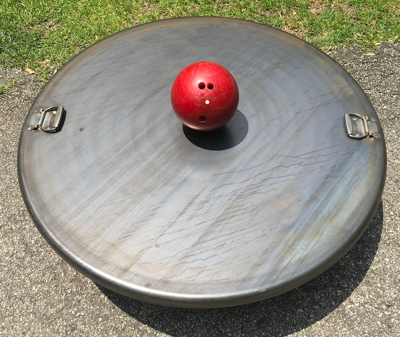 May include: A red bowling ball sits on a large, round, metal disc with two metal handles. The disc has a textured surface.