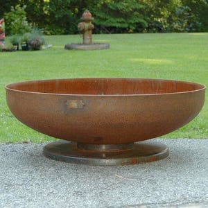 Op de afbeelding: Een grote, roestige metalen vuurplaats met een brede, ronde kom en een verhoogde basis. De vuurplaats staat op een grijs, geplaveid oppervlak.