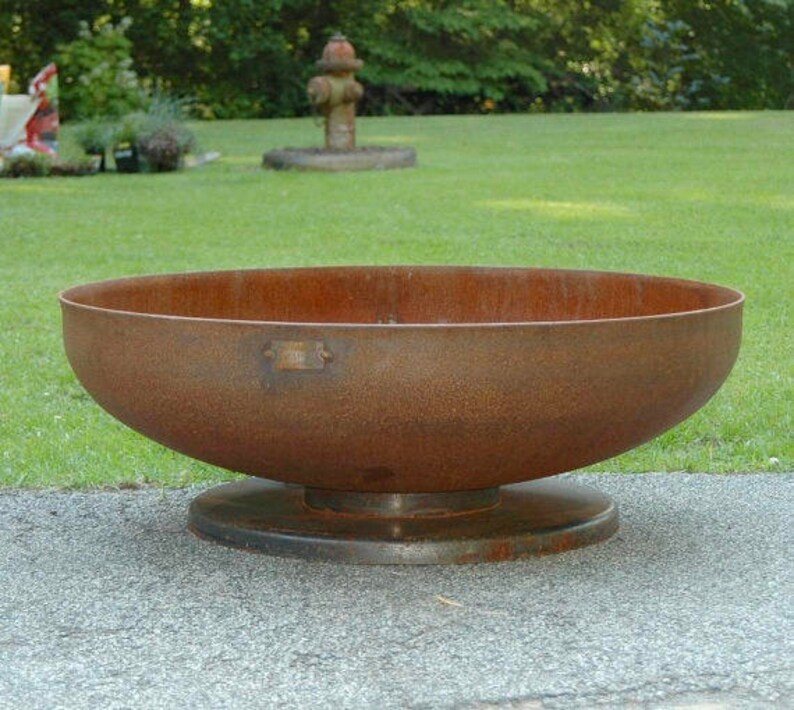 Op de afbeelding: Een grote, roestige metalen vuurplaats met een brede, ronde kom en een verhoogde basis. De vuurplaats staat op een grijs, geplaveid oppervlak.