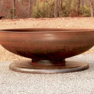 May include: A large, circular, bowl-shaped fire pit made of weathered metal with a distinct rusted brown patina. The fire pit features a smooth, curved basin that rests securely on a flat, circular base, creating a low-profile, stable structure.