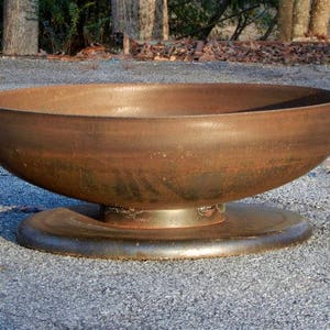 May include: A large, circular, bowl-shaped fire pit made of rusted metal. The basin features a smooth, weathered brown patina and is supported by a sturdy, flat, circular metal base that provides stability for the structure on the ground.