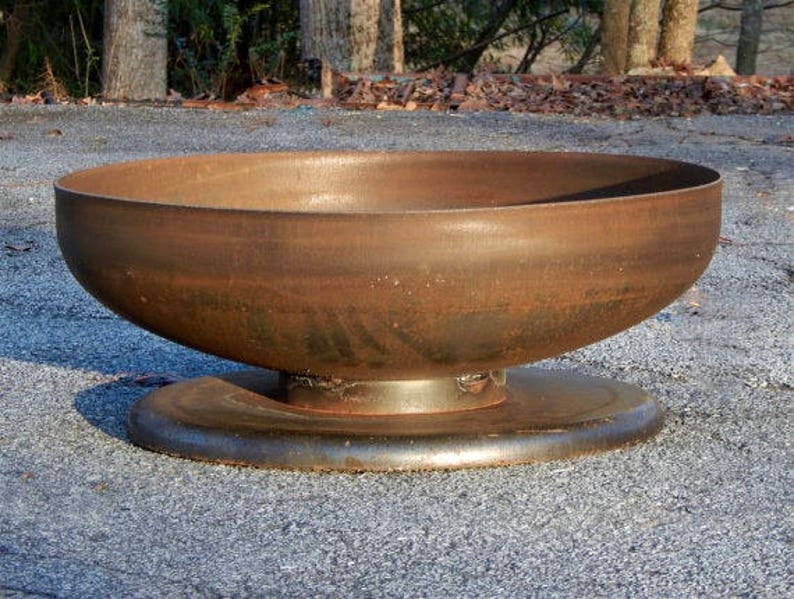 May include: A large, circular, bowl-shaped fire pit made of rusted metal. The basin features a smooth, weathered brown patina and is supported by a sturdy, flat, circular metal base that provides stability for the structure on the ground.