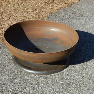 May include: A circular metal fire pit bowl with a weathered, rusted brown finish. The bowl features a smooth, curved interior and exterior surface, resting on a solid, dark-colored circular base. A small drainage hole is visible at the bottom.