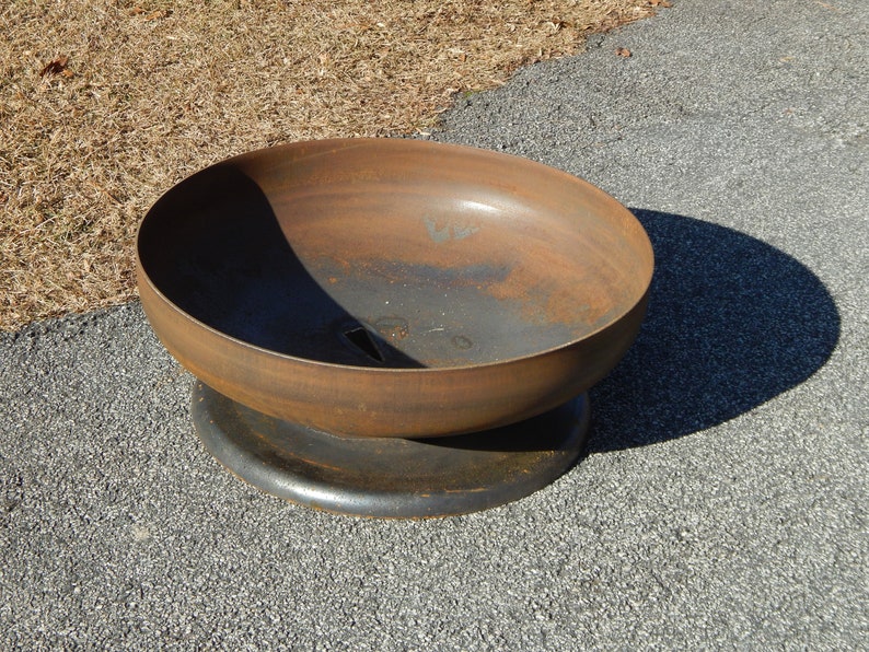 May include: A circular metal fire pit bowl with a weathered, rusted brown finish. The bowl features a smooth, curved interior and exterior surface, resting on a solid, dark-colored circular base. A small drainage hole is visible at the bottom.