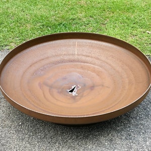 May include: A large, rusty metal fire pit with a circular shape and a small hole in the center. The fire pit is sitting on a gray asphalt surface.