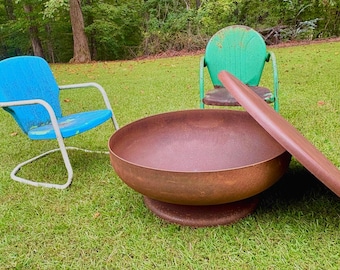 May include: A large, round, rusted metal fire pit bowl with a matching circular lid resting beside it. To the left is a blue metal chair with a white frame, and behind the fire pit is a green metal chair with a weathered, rusted finish.