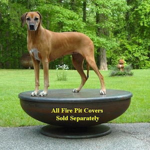 Op de afbeelding: Een bruine hond staat op een grote, ronde, metalen vuurplaatsdeksel. Het deksel staat op een voetstuk. De tekst "All Fire Pit Covers Sold Separately" staat op het deksel gedrukt.