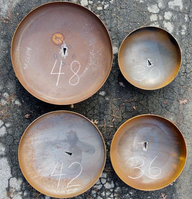 Op de afbeelding: Vier ronde metalen vuurkorven in verschillende maten. De kommen zijn roestbruin en hebben witte nummers erop geschreven. De nummers zijn 48, 30, 42 en 36.
