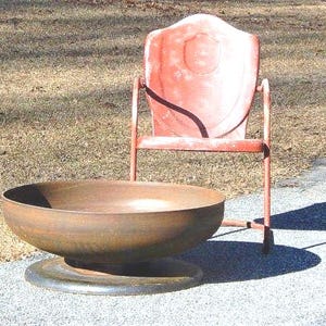 May include: A large, circular, rusted metal fire pit bowl with a wide, flat base, positioned in front of a vintage, weathered red metal chair with a shield-shaped backrest and thin tubular legs, both resting on a paved surface.