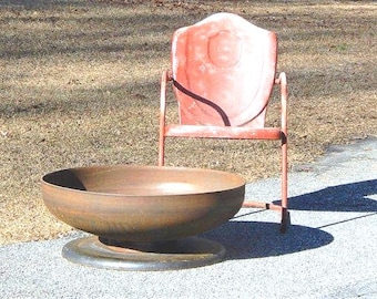 May include: A large, circular, rusted metal fire pit bowl with a wide, flat base, positioned in front of a vintage, weathered red metal chair with a shield-shaped backrest and thin tubular legs, both resting on a paved surface.