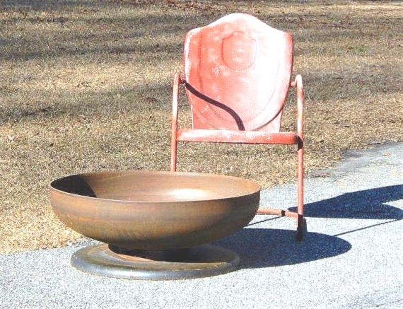 May include: A large, circular, rusted metal fire pit bowl with a wide, flat base, positioned in front of a vintage, weathered red metal chair with a shield-shaped backrest and thin tubular legs, both resting on a paved surface.