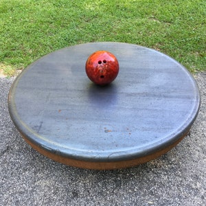 May include: A red bowling ball with three holes sits on a round, gray, textured surface.