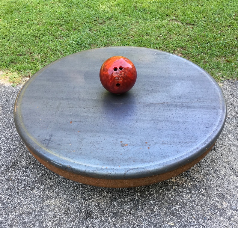 May include: A red bowling ball with three holes sits on a round, gray, textured surface.