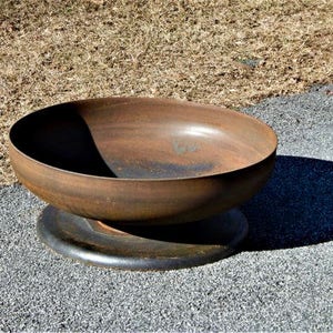 May include: A circular, bowl-shaped fire pit made of weathered, rusted metal with a smooth, deep basin. The fire pit rests on a flat, dark, circular metal base, featuring a uniform, oxidized brown patina across its exterior and interior surfaces.