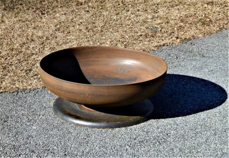 May include: A circular, bowl-shaped fire pit made of weathered, rusted metal with a smooth, deep basin. The fire pit rests on a flat, dark, circular metal base, featuring a uniform, oxidized brown patina across its exterior and interior surfaces.