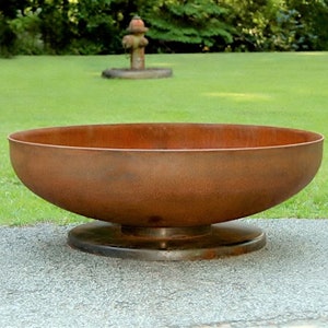 Op de afbeelding: Een grote, roestige metalen vuurkorf met een brede, ondiepe basis en een verhoogde rand. De kom staat op een platte, ronde basis.