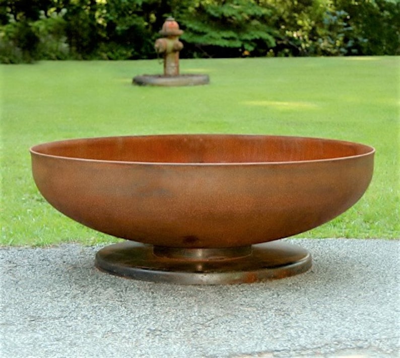Op de afbeelding: Een grote, roestige metalen vuurkorf met een brede, ondiepe basis en een verhoogde rand. De kom staat op een platte, ronde basis.