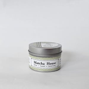 May include: A round silver metal tin with a lid, featuring a label with a light green geometric border. The label displays the text Matcha House, Bitter + Sweet + Ceremony in black font. The lid also has a small circular label with text.