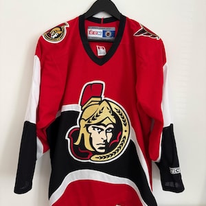 May include: A red, black, and white hockey jersey with the Ottawa Senators logo on the front. The jersey has a V-neck and white sleeves with black cuffs. The team logo features a Roman soldier's head.