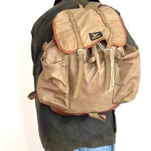 May include: A tan backpack with brown leather trim and multiple pockets. The backpack has adjustable straps and a flap closure with buckles. A small patch with a logo is visible on the front. The backpack is worn by a person wearing a green jacket and blue jeans.