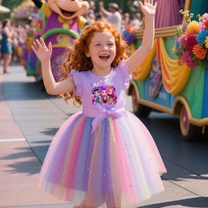 Peut inclure: Une jeune fille rousse sourit et salue, vêtue d'une robe lavande avec une jupe en tulle arc-en-ciel. La robe a un nœud et une impression graphique. Un char de défilé et des personnes sont visibles en arrière-plan.