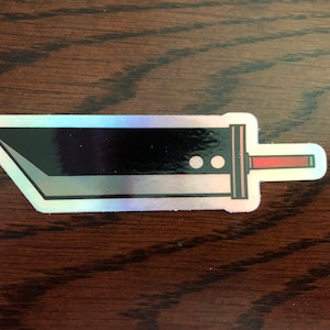 May include: A holographic sticker of a black sword with a red handle. The sticker is on a brown wooden surface.