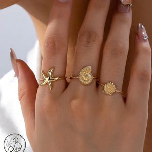 May include: Three gold-colored rings with ocean-themed designs are displayed on a hand. The rings feature a starfish, a seashell, and a scallop shell. The rings are delicate and have a polished finish, suggesting they are made of metal.