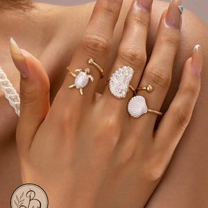 May include: A set of three gold-toned rings worn on fingers. One ring features a white turtle, another displays an irregular white textured piece, and the third ring showcases a white fan-shaped seashell design with a small gold bead at the end.