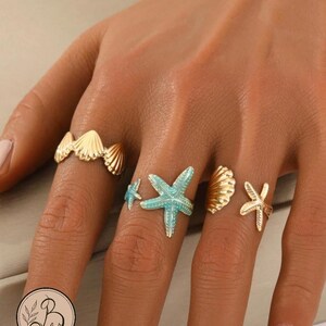 May include: A set of four gold-toned rings featuring coastal motifs. One ring displays a row of small seashells, another features a large turquoise starfish with a smaller matching starfish, and the final two rings show a single seashell and starfish.