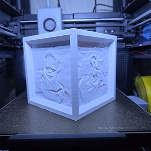 May include: A white, three-dimensional cube with relief portraits on each side, likely created using a 3D printer. The cube is positioned on a black surface within a 3D printer, showcasing the technology's capabilities. The portraits appear to be of a person.
