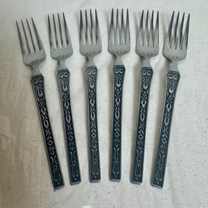 May include: A set of six silver-toned metal forks arranged in a row. Each fork features four tines and a long, rectangular handle decorated with a dark, recessed, repeating scrollwork pattern that extends from the neck to the base of the handle.