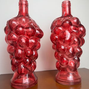 May include: Two red glass bottles with a grape cluster design. The bottles have a bulbous shape and a long neck. The bottles are made of transparent red glass and are sitting on a brown surface.