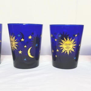 May include: Four cobalt blue drinking glasses with celestial designs. Each glass features a yellow sun, crescent moon, and stars. The glasses have a gradient effect, transitioning from dark blue to a lighter shade.