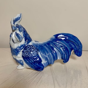 May include: A ceramic rooster figurine featuring a white base with intricate blue hand-painted patterns. The design showcases detailed plumage on the wings and tail, a prominent comb on the head, and a glossy finish throughout the entire sculpture.