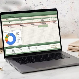 May include: A laptop displaying a financial planning spreadsheet with a pie chart, next to straw hats, a paperclip, earrings, and folded tan fabric. A notebook and pen are also visible.