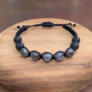 May include: A bracelet featuring a black cord with an adjustable sliding knot closure. The design includes five smooth, translucent gray spherical beads in the center, flanked by four textured black lava stone beads and two small silver-toned metal beads.
