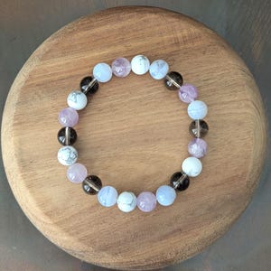 May include: A circular beaded bracelet featuring a repeating pattern of smooth, round beads in shades of translucent light blue, opaque white with grey veining, pale lavender, and dark translucent brown, all strung together on an elastic cord.