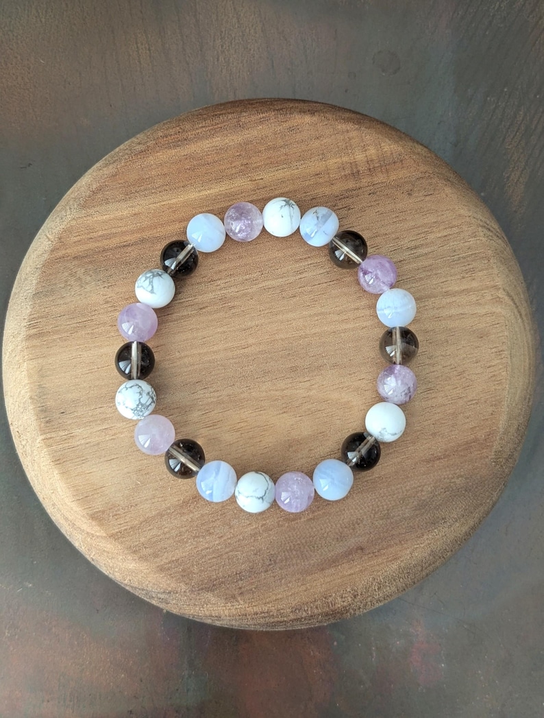 May include: A circular beaded bracelet featuring a repeating pattern of smooth, round beads in shades of translucent light blue, opaque white with grey veining, pale lavender, and dark translucent brown, all strung together on an elastic cord.