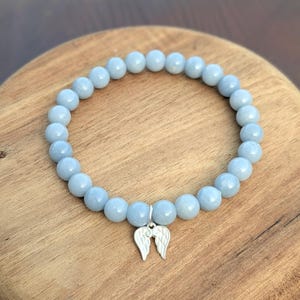 Puede incluir: Una pulsera de cuentas azul claro con un dije de alas de &aacute;ngel plateadas. La pulsera est&aacute; hecha de cuentas redondas y lisas y se exhibe sobre una superficie de madera. Es una joya.