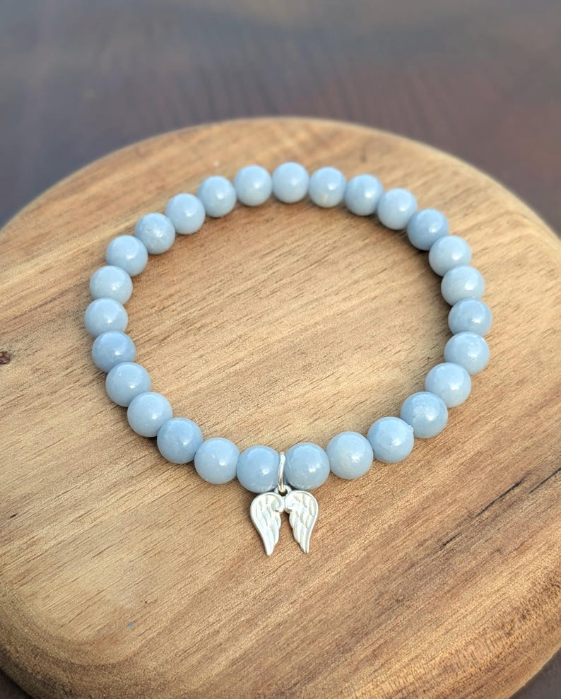 Puede incluir: Una pulsera de cuentas azul claro con un dije de alas de &aacute;ngel plateadas. La pulsera est&aacute; hecha de cuentas redondas y lisas y se exhibe sobre una superficie de madera. Es una joya.