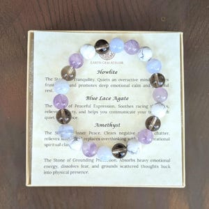 May include: A circular beaded bracelet featuring alternating smooth round stones in white, translucent light blue, dark brown, and light purple. The bracelet rests on a square card printed with the text Earth Gem Atelier, Howlite, Blue Lace Agate, and Amethyst, along with descriptive paragraphs about each stone type.