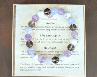 May include: A circular beaded bracelet featuring alternating smooth round stones in white, translucent light blue, dark brown, and light purple. The bracelet rests on a square card printed with the text Earth Gem Atelier, Howlite, Blue Lace Agate, and Amethyst, along with descriptive paragraphs about each stone type.
