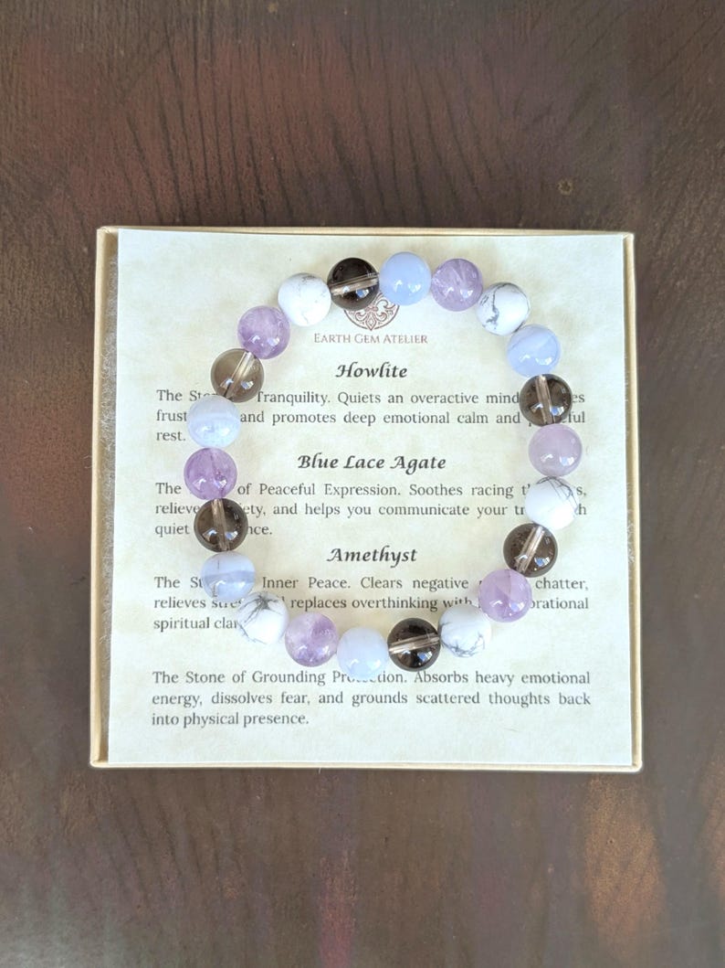 May include: A circular beaded bracelet featuring alternating smooth round stones in white, translucent light blue, dark brown, and light purple. The bracelet rests on a square card printed with the text Earth Gem Atelier, Howlite, Blue Lace Agate, and Amethyst, along with descriptive paragraphs about each stone type.