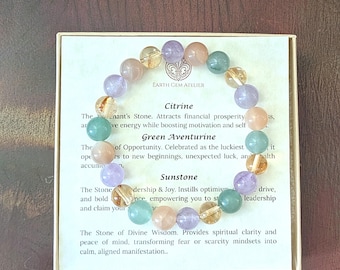 May include: A circular beaded bracelet featuring polished spherical stones in shades of purple, translucent yellow, peach, and green. The bracelet rests on a square card printed with the text Earth Gem Atelier, Citrine, Green Aventurine, and Sunstone.