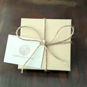 May include: A square, light brown cardboard gift box tied with thin, natural-colored jute twine in a bow. A small, rectangular white card is tucked under the twine, featuring a circular logo with a fleur-de-lis and the text EARTH GEM ATELIER.