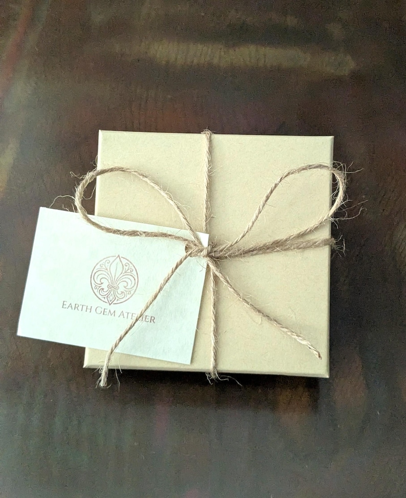 May include: A square, light brown cardboard gift box tied with thin, natural-colored jute twine in a bow. A small, rectangular white card is tucked under the twine, featuring a circular logo with a fleur-de-lis and the text EARTH GEM ATELIER.