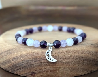 May include: A beaded bracelet featuring alternating dark purple and light lavender spherical beads. The bracelet includes a silver-toned metal charm shaped like a crescent moon with an intricate filigree cutout pattern hanging from the center.