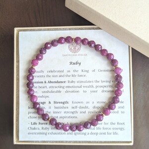 May include: A circular beaded bracelet made of faceted, deep pink ruby gemstones with small gold-toned spacers. The bracelet rests on a square card inside an open box, featuring the text Earth Gem Atelier and Ruby with descriptive text.