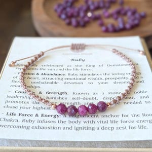 May include: A delicate adjustable bracelet featuring five faceted deep pink ruby beads centered on a light brown knotted cord. The bracelet rests on a white card printed with text about ruby gemstones, with a small pile of loose ruby beads visible.