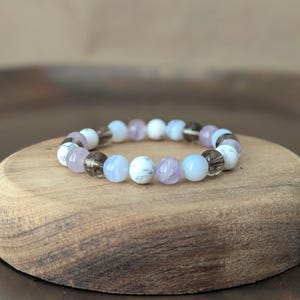 May include: A beaded bracelet featuring a repeating pattern of round, polished stones in shades of white with grey veining, translucent light blue, pale lavender, and dark smoky brown, all strung together on a flexible, circular elastic cord.